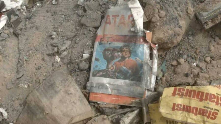 Dettaglio dei rifiuti rinvenuti nello scavo: una confezione in cartone del videogioco E.T. l'Extra-Terrestre per Atari, visibilmente schiacciata, sgualcita e ricoperta di polvere, giace tra terra e detriti.