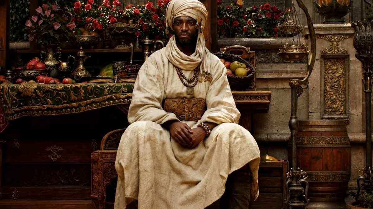 Un personaggio maschile indossa un turbante e vesti chiare ed è seduto in un mercato estremamente dettagliato, circondato da cesti di frutta, vasi finemente decorati e fiori rossi.