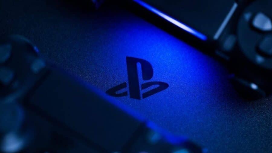 Dettaglio ravvicinato del logo PlayStation inciso su una console scura, illuminato da una suggestiva luce blu neon, con parti di un joypad sfocate in primo piano.