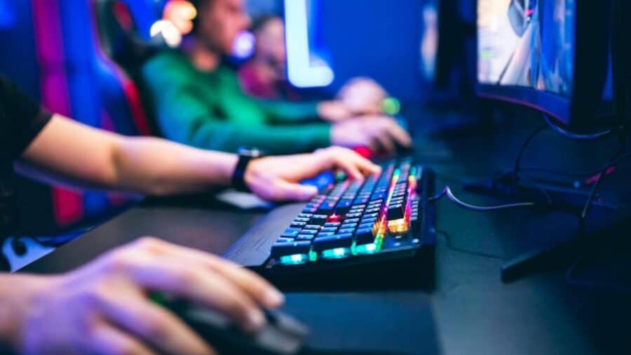Primo piano delle mani di un videogiocatore appoggiate su una tastiera meccanica retroilluminata RGB, con altri gamer sfocati sullo sfondo durante una sessione di gioco competitiva.
