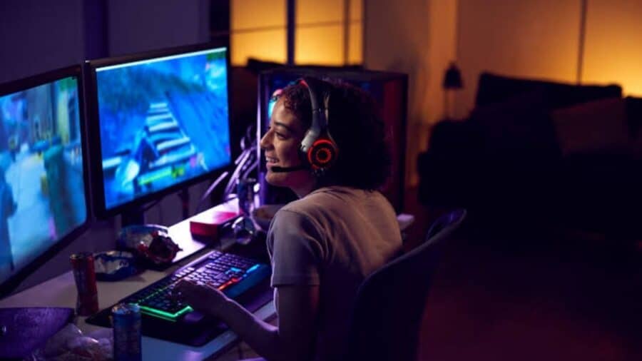 Una ragazza sorridente indossa delle cuffie da gaming con luci rosse mentre gioca al PC davanti a un doppio monitor in una stanza in penombra.