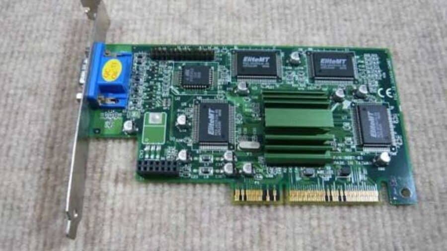 Una scheda grafica vintage con circuito stampato verde, chip di memoria e un dissipatore di calore alettato in metallo verde, poggiata su una superficie grigia.