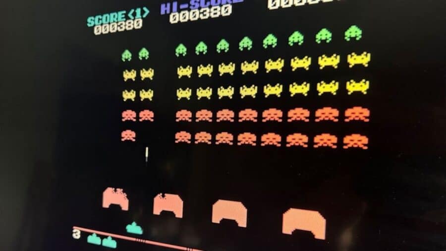 Dettaglio ravvicinato dello schermo di un cabinato arcade di Space Invaders, che mostra i classici alieni pixelati divisi in file di colore verde, giallo e rosso che scendono verso i bunker difensivi.