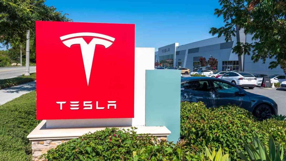 Una grande insegna rossa con il logo bianco e la scritta Tesla, posizionata all'esterno di un concessionario con diverse auto elettriche parcheggiate sullo sfondo.