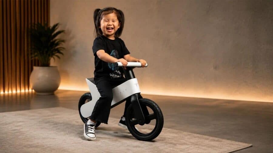 Una bambina sorridente e felice mentre spinge con i piedi la balance bike bianca targata Tesla all'interno di un salotto dal design moderno e minimale.