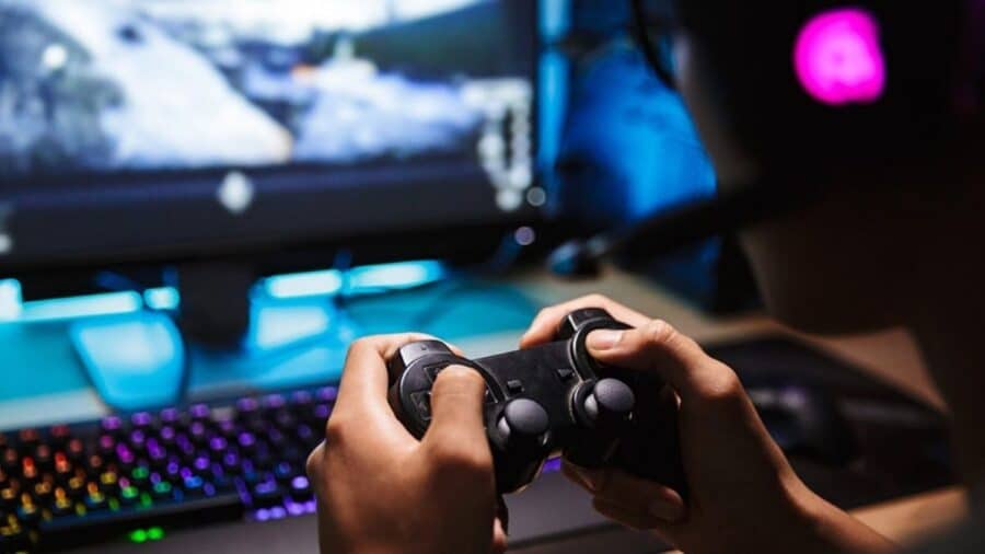 Dettaglio ravvicinato delle mani di un videogiocatore che impugna un controller nero; sullo sfondo sfocato si intravedono un monitor acceso e una tastiera da gaming illuminata con LED RGB.