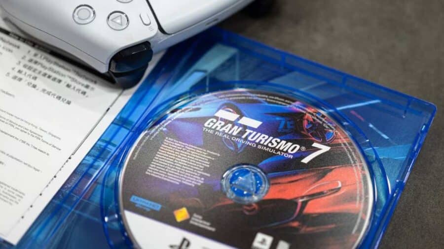 Custodia aperta del videogioco Gran Turismo 7 per PlayStation con il disco ottico in primo piano, affiancata da un controller DualSense bianco e da un foglietto di istruzioni cartaceo.