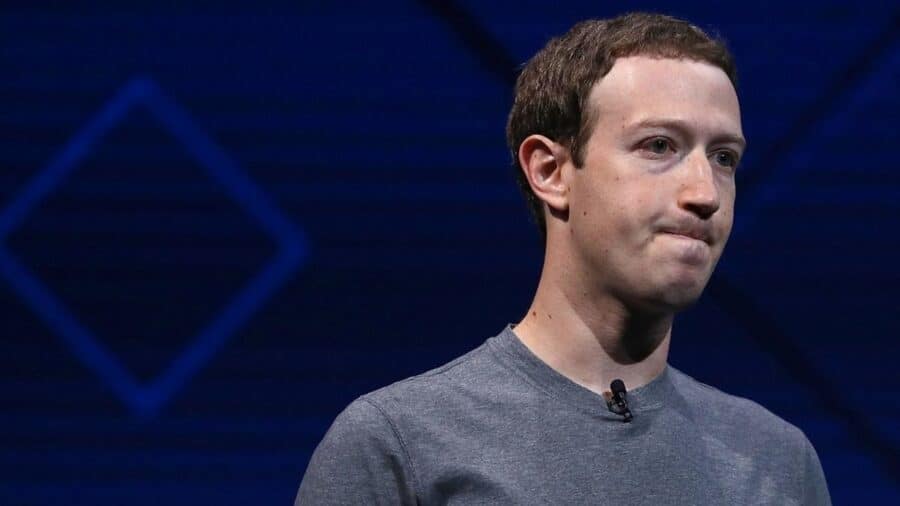 Mezzo busto di Mark Zuckerberg con un'espressione seria e le labbra serrate, mentre indossa la sua iconica t-shirt grigia su uno sfondo blu geometrico.