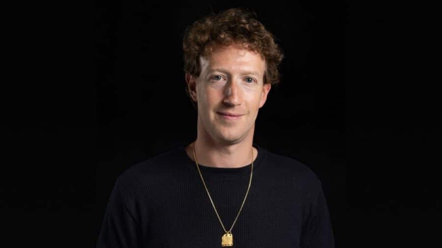 Ritratto in primo piano di Mark Zuckerberg con un look recente, caratterizzato da capelli mossi e una catenina d'oro al collo, che sorride leggermente su uno sfondo completamente nero.