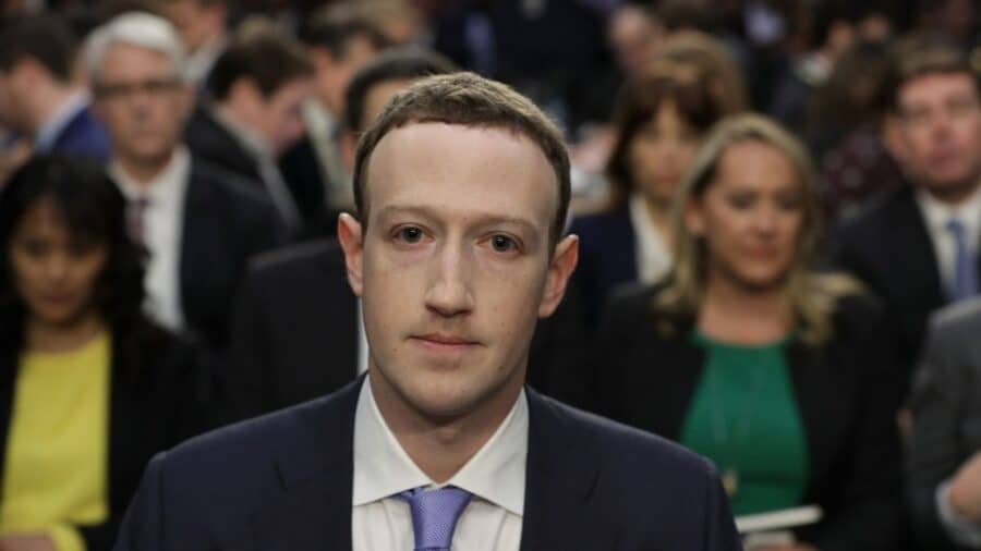 Mark Zuckerberg indossa un abito formale scuro e una cravatta azzurra, con uno sguardo teso e concentrato, seduto in un'aula con altre persone sfocate sullo sfondo.