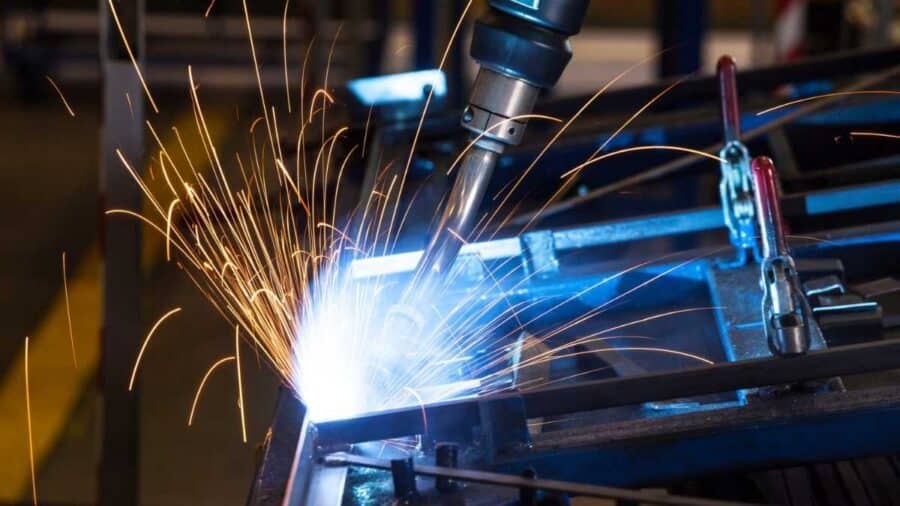 Dettaglio ravvicinato di un braccio robotico industriale in piena azione durante un'operazione di saldatura su una griglia metallica, circondato da un'intensa pioggia di scintille luminose.