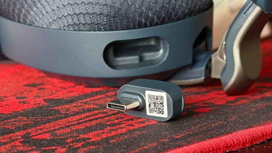 Dongle wireless USB-C delle cuffie MSI Maestro 500 appoggiato su un tappetino per mouse rosso e nero, con il padiglione delle cuffie visibile in parte sullo sfondo.