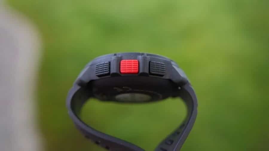 Dettaglio del profilo laterale dello smartwatch Polar Street X su sfondo verde sfocato, che mostra la spessa cassa grigia e tre pulsanti fisici testurizzati, con il tasto centrale in forte contrasto di colore rosso.