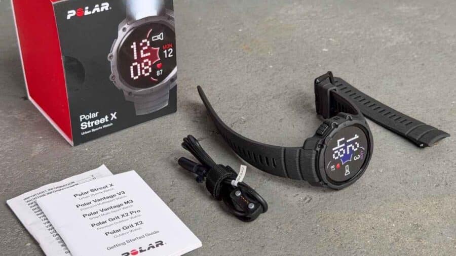 Contenuto della confezione del Polar Street X appoggiato su un piano grigio: in evidenza lo smartwatch scuro, la scatola originale bianca e rossa, il cavo di ricarica USB proprietario e i manuali di istruzioni.