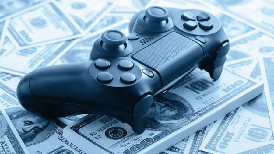 Un controller da gioco nero di vecchia generazione appoggiato sopra una distesa di banconote da cento dollari statunitensi, caratterizzato da un filtro fotografico sui toni dell'azzurro.