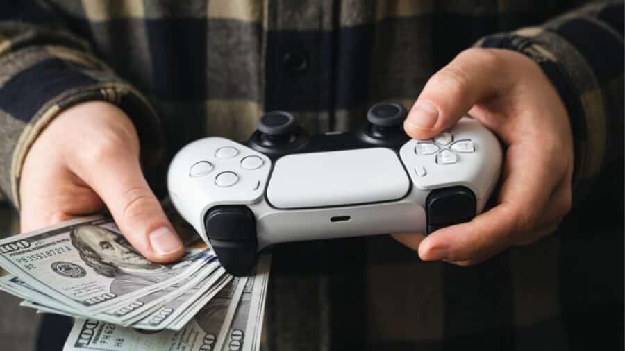 Primo piano delle mani di un giocatore che stringono contemporaneamente un moderno controller bianco per PlayStation 5 (DualSense) e un corposo mazzetto di banconote da cento dollari.