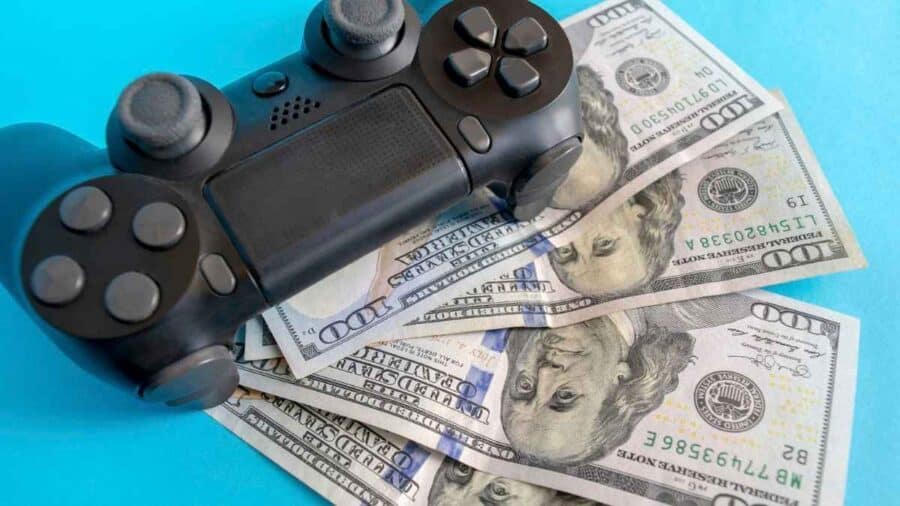 Controller nero in stile PlayStation posato su alcune banconote da cento dollari sparpagliate, in forte contrasto con uno sfondo azzurro acceso.