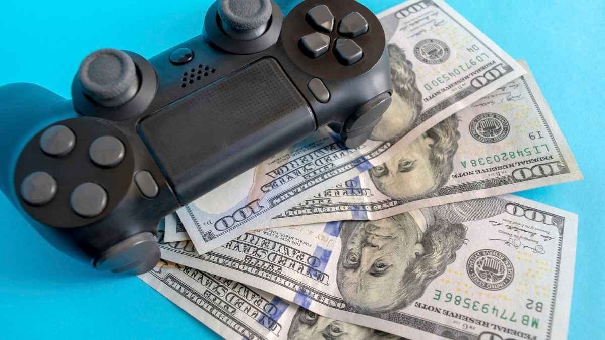 Controller nero in stile PlayStation posato su alcune banconote da cento dollari sparpagliate, in forte contrasto con uno sfondo azzurro acceso.