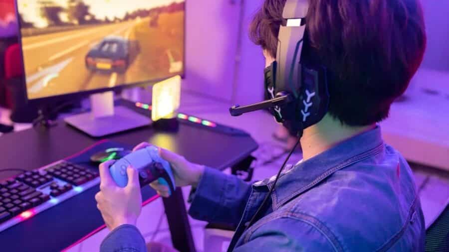Un ragazzo ripreso di spalle mentre gioca a un simulatore di guida davanti a un monitor; indossa vistose cuffie da gaming, impugna un controller e la stanza è illuminata da soffuse luci LED viola e blu.