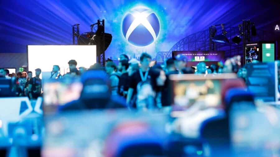 Folla di videogiocatori sfocata in primo piano durante una fiera di settore, sovrastata da un grande e luminoso logo Xbox azzurro e bianco.