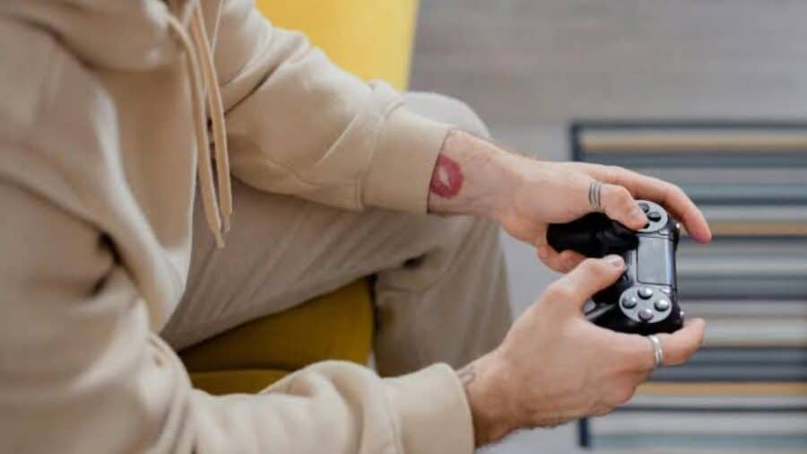 Dettaglio ravvicinato delle mani di un videogiocatore, con anelli e piccoli tatuaggi sulle dita, che impugna un controller nero per PlayStation (DualShock 4) mentre è seduto su un divano.