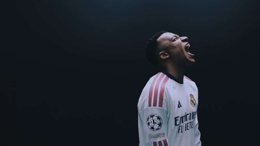 Primo piano cinematografico di Kylian Mbappé con la nuova maglia bianca del Real Madrid, che urla a occhi chiusi in un'intensa esultanza su uno sfondo completamente scuro.