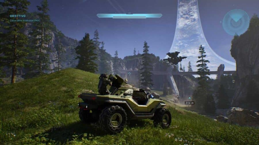 Visuale in terza persona di Master Chief alla guida di un fuoristrada Warthog verde in un paesaggio boschivo, mentre la maestosa struttura ad anello di Halo si staglia nel cielo curvando verso l'alto.