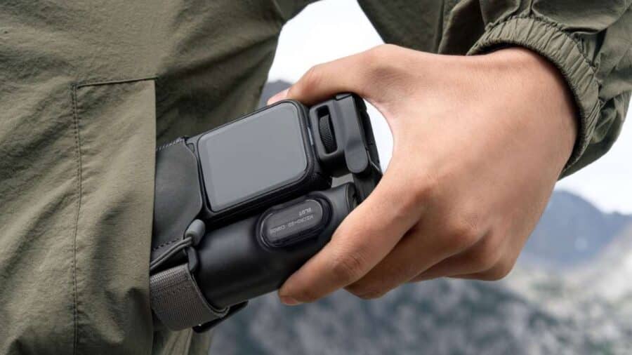 Primo piano della mano di una persona che estrae la nuova videocamera Insta360 X6 dalla tasca di una giacca a vento verde. È visibile il grande schermo OLED da 2,32 pollici e lo sportellino laterale per la porta USB e la Micro-SD.
