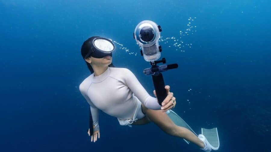 Una subacquea in muta bianca nuota sott'acqua nel blu profondo dell'oceano, tenendo in mano una Insta360 X6 racchiusa all'interno dello speciale scafandro trasparente Invisible Dive Case Pro.