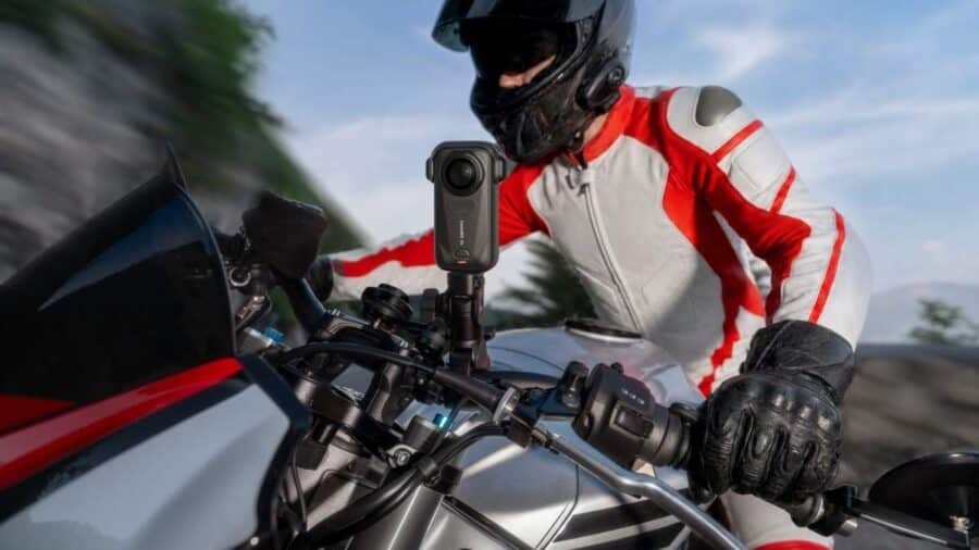 Inquadratura dinamica dal punto di vista del manubrio di una moto sportiva in corsa. In primo piano, la videocamera Insta360 X6 è saldamente ancorata al telaio mentre il motociclista in tuta bianca e rossa piega in curva.