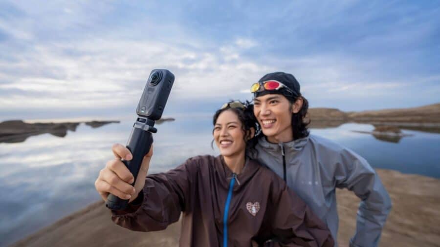 Una giovane coppia sorridente si scatta un selfie panoramico utilizzando la videocamera Insta360 X6 montata su un bastone per selfie (Selfie Stick). Sullo sfondo, un lago calmo riflette le formazioni rocciose sotto un cielo parzialmente nuvoloso.