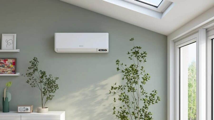 Un moderno condizionatore split bianco installato sulla parete verde salvia di un soggiorno luminoso, sopra un mobiletto con una pianta d'appartamento.