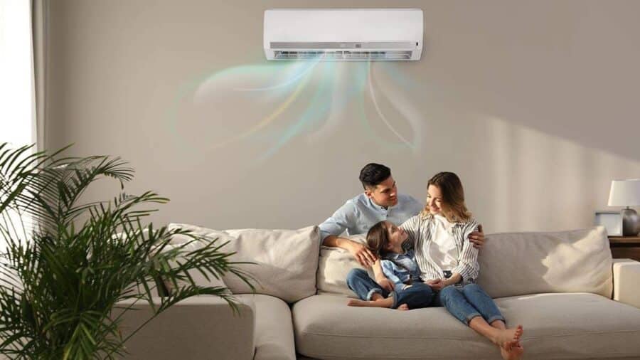 Una famiglia felice, composta da madre, padre e una bambina, sorride abbracciata su un comodo divano beige nel soggiorno di casa, mentre un condizionatore a parete diffonde aria fresca stilizzata con sfumature arcobaleno.