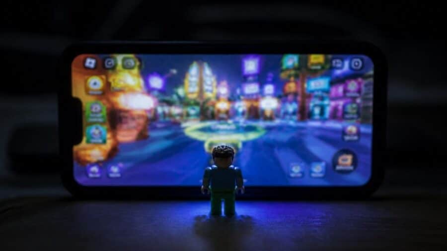 Una piccola figura giocattolo inquadrata di spalle osserva lo schermo luminoso di uno smartphone al buio, sul quale è in esecuzione una colorata mappa virtuale del videogioco Roblox.