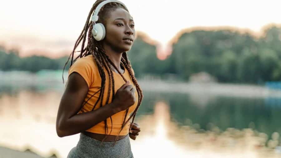 Una ragazza con le treccine indossa delle cuffie bianche over-ear e abbigliamento sportivo giallo e grigio, concentrata mentre corre all'aperto vicino a uno specchio d'acqua al tramonto.