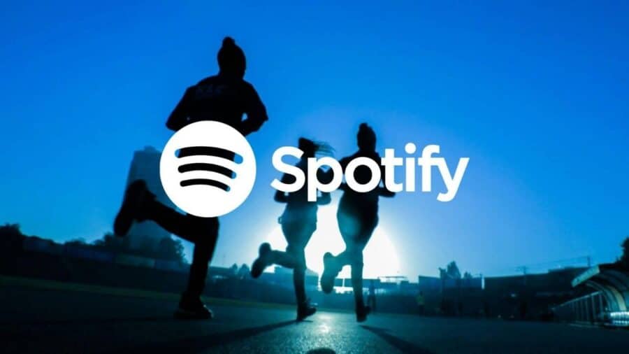 Il classico logo bianco di Spotify sovrapposto alla silhouette in controluce di tre persone che corrono insieme su una pista sportiva, con un cielo azzurro brillante e il sole basso sull'orizzonte.