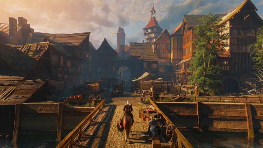 Geralt di Rivia a cavallo di Rutilia attraversa un ponte di legno verso l'ingresso di un vivace borgo medievale con case a graticcio e una torre campanaria al tramonto.