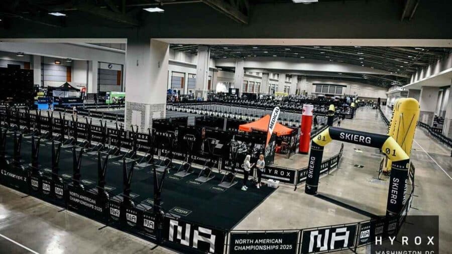 Panoramica dell'arena interna di una competizione HYROX con la stazione dello SkiErg contrassegnata da un grande arco gonfiabile giallo e nero davanti a file di simulatori Concept2.
