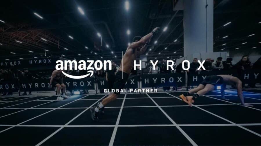 Grafica ufficiale con i loghi Amazon e HYROX Global Partner in primo piano su uno scatto di atleti che compiono burpee broad jump su un tracciato a griglia.