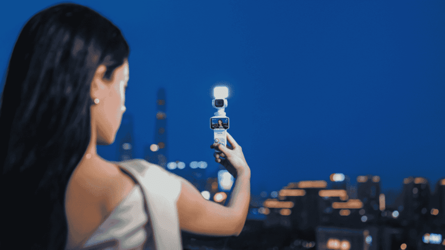 Donna di spalle che impugna la videocamera gimbal bianca Insta360 Luna Pro con faretto ausiliario acceso, registrando un vlog notturno con le luci dello skyline cittadino sfocate sullo sfondo.