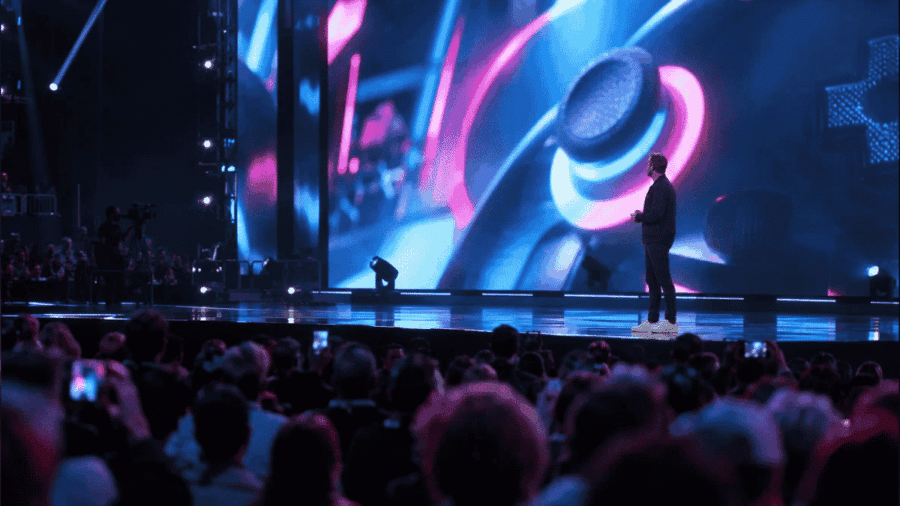 Una vista panoramica dal basso di un grande palcoscenico per conferenze, con un presentatore in piedi davanti a un enorme schermo LED curvo che mostra grafiche di gioco luminose in blu e rosa, e un pubblico numeroso che osserva.