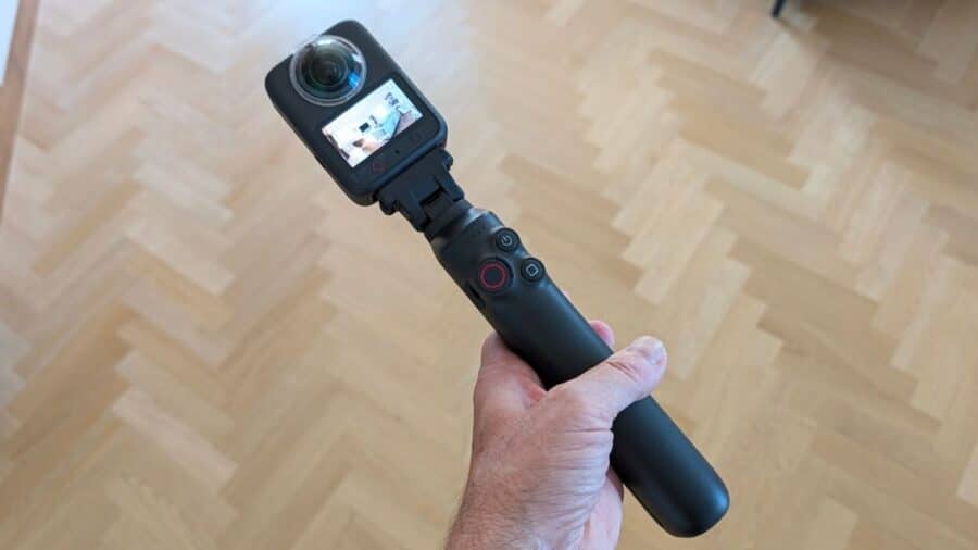 Mano che regge Osmo 360 II montata su un bastone selfie invisibile, con pavimento in parquet a spina di pesce sullo sfondo e schermo della videocamera acceso.