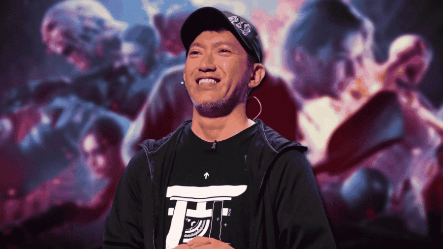 Primo piano di Shinji Mikami sorridente sul palco con un cappellino nero con visiera, maglietta scura e microfono ad archetto, con una grafica di gioco sullo sfondo.
