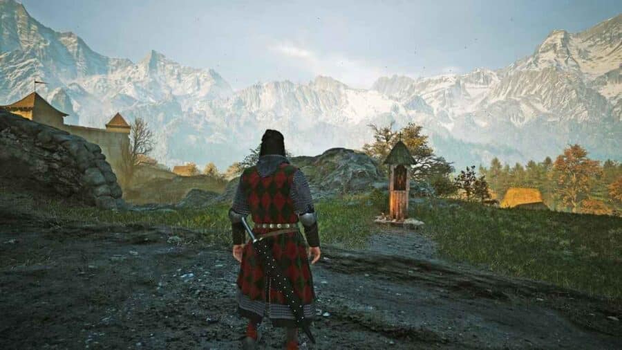 Coen visto di spalle con una tunica a rombi rossi e verdi mentre osserva un piccolo santuario di legno illuminato lungo un sentiero montano, sovrastato dalle vette innevate dei Carpazi.
